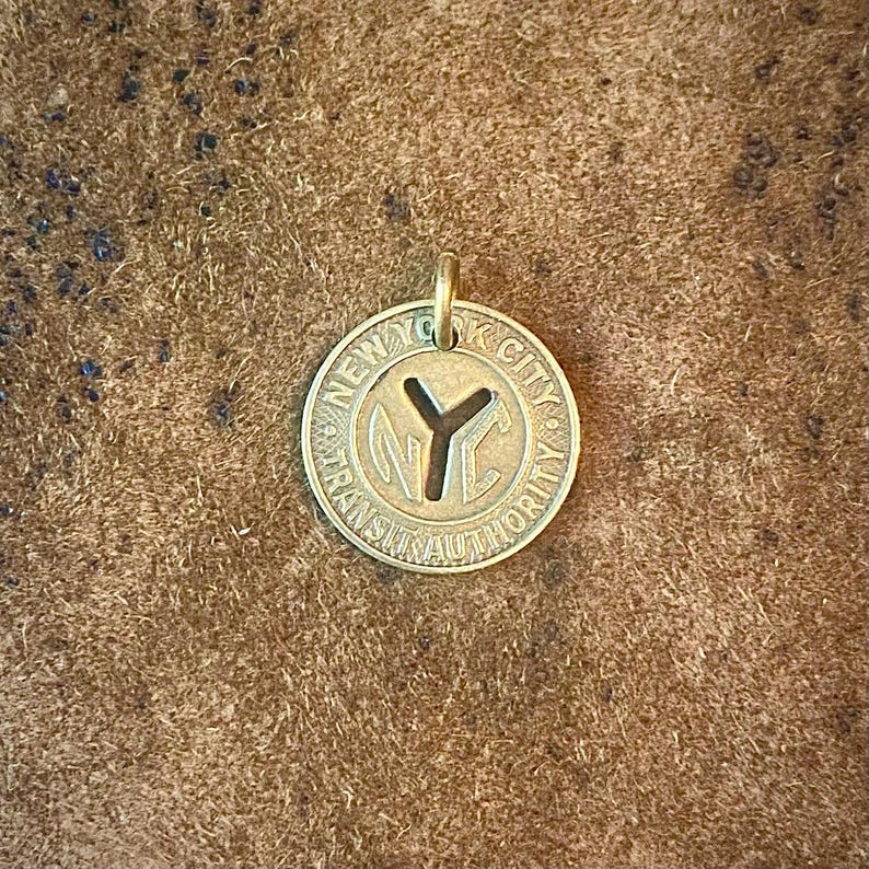 May include: A circular brass pendant featuring the text "NEW YORK CITY TRANSIT AUTHORITY" around the edge. The center has a cutout of the letters "NYC" and a "Y" shape. The pendant has a small loop at the top for a chain.
