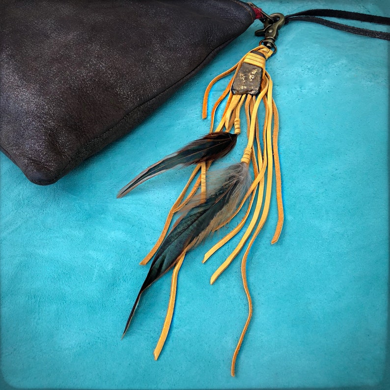 May include: A brown leather bag with a long tassel attached. The tassel is made of leather and feathers and has a gold metal charm.