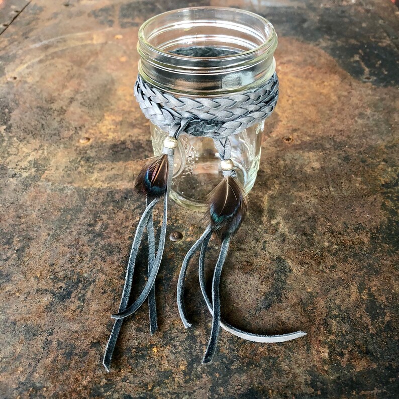 May include: A glass jar with a gray braided leather band and two gray leather tassels with black feathers. The jar is sitting on a rustic metal surface.
