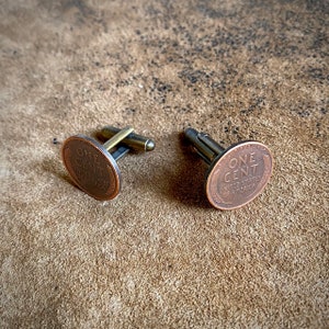 May include: A pair of cufflinks featuring a copper-toned oval design with the words "ONE CENT" and "UNITED STATES OF AMERICA".