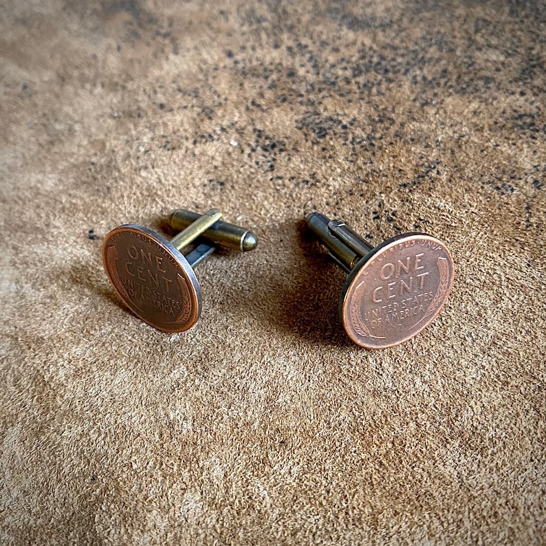 May include: A pair of cufflinks featuring a copper-toned oval design with the words "ONE CENT" and "UNITED STATES OF AMERICA".