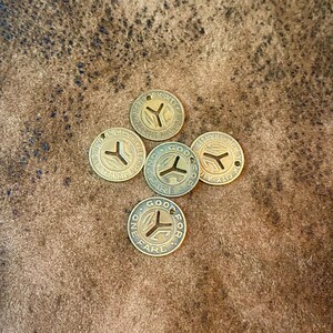 May include: Five antique brass transit tokens with a Y-shaped design and the words "GOOD FOR FARE" and "NEW YORK CITY" embossed around the edges. Each token has a small hole at the top. The tokens are arranged on a brown, textured surface.