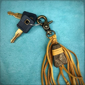 May include: A gold key chain with a brown leather tassel and a small, dark brown stone charm. The keychain is attached to two keys with black plastic handles.