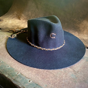 May include: A black felt cowboy hat with a brown leather braided band and a horseshoe logo. The hat has a wide brim and a tall crown.