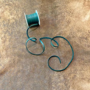 May include: A spool of dark teal leather cord with a length of the cord unraveled and curled on a brown surface. The cord is thin and appears to be made of a smooth material. The spool is white.