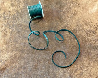 May include: A spool of dark teal leather cord with a length of the cord unraveled and curled on a brown surface. The cord is thin and appears to be made of a smooth material. The spool is white.