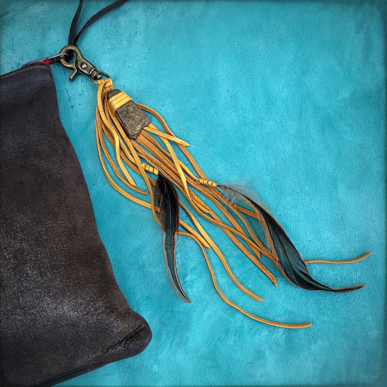 May include: A brown leather bag with a long, yellow tassel attached. The tassel has a small, dark brown stone at the top and black feathers hanging from the bottom. The bag and tassel are on a turquoise background.