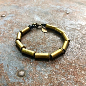 May include: A gold-toned bracelet made of bullet casings. The bracelet has a black cord closure.