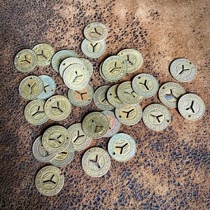 May include: A collection of vintage New York City Transit Authority tokens. The brass-colored tokens are circular with a stylized "Y" design and the text "Good for One Fare". The tokens are scattered on a textured brown surface.
