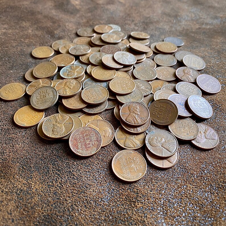 May include: A large pile of copper-colored US one-cent coins, also known as pennies, on a brown leather surface. The coins are arranged in a random pattern, with some coins overlapping others. The coins are all in good condition, with no visible damage or wear.