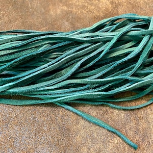 May include: Close-up of a bundle of teal-colored leather strips. The strips are thin, flexible, and have a slightly textured surface. The leather is a vibrant teal color, with variations in tone. The background is a brown, textured surface.