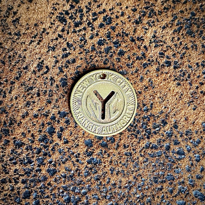 May include: A vintage brass New York City Transit Authority token. The token is circular with the text "NEW YORK CITY TRANSIT AUTHORITY" around the edge and a "Y" symbol in the center. The token has a small hole at the top.