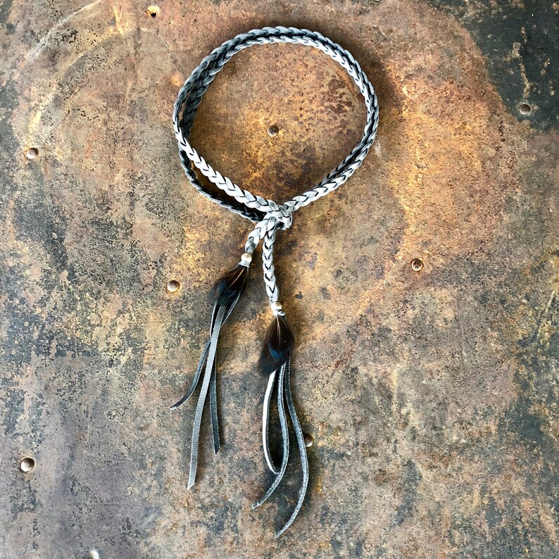 May include: A braided leather cord with black feather tassels. The cord is gray and white and is tied in a knot at the top. The tassels are long and flowing.