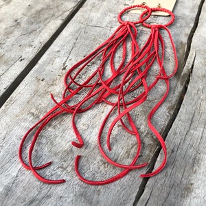 May include: A pair of red leather earrings with long fringe tassels. The earrings are made of thin leather strips that are tied together at the top. The fringe is long and flowing, and the earrings are a bright red colour.