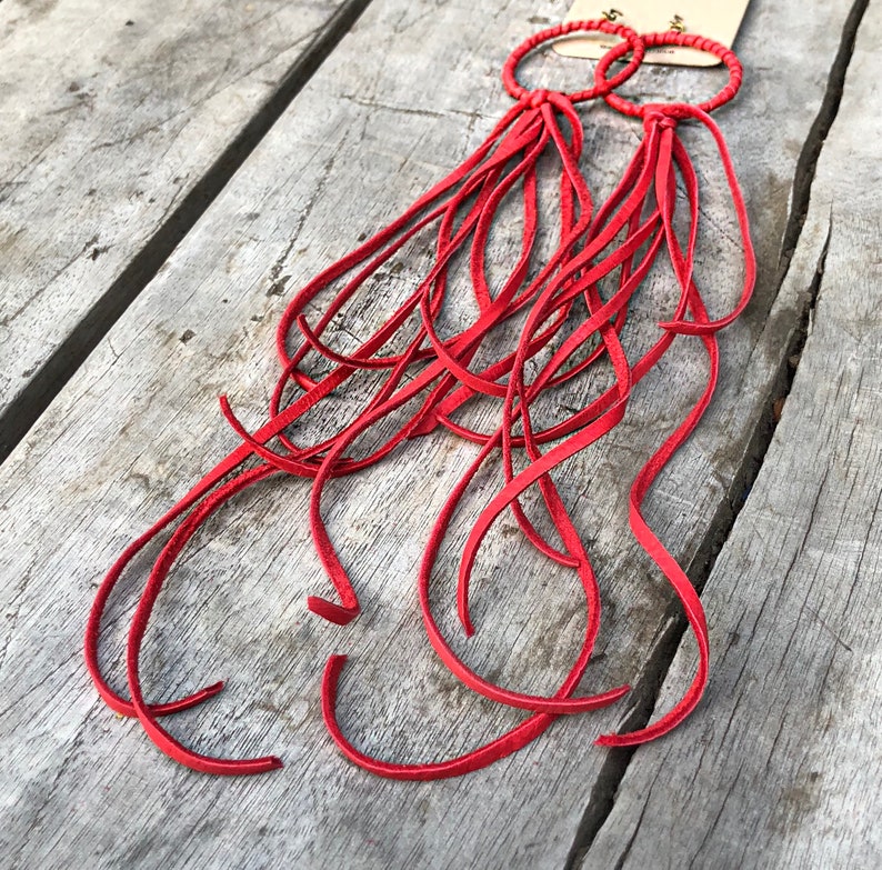 May include: A pair of red leather earrings with long fringe tassels. The earrings are made of thin leather strips that are tied together at the top. The fringe is long and flowing, and the earrings are a bright red colour.