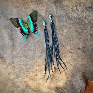 May include: A pair of long, dark blue feather earrings with turquoise accents and gold-colored hooks. A butterfly with green, blue, and brown wings is also present. The earrings are displayed on a textured, brown surface.