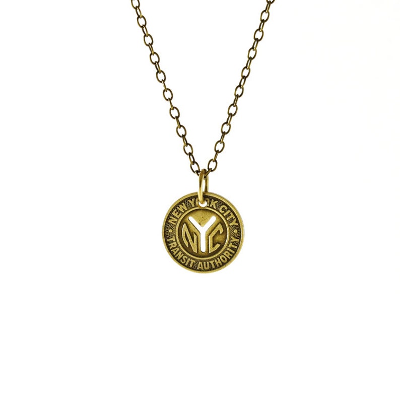 May include: A gold-colored necklace with a round pendant. The pendant features the text "NYC TRANSIT AUTHORITY" around the edge and the letters "NYC" in the center. The chain is a matching gold color.