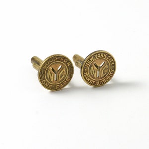 May include: Two vintage brass cufflinks. Each cufflink is round with the letters "NYC" in the center. One cufflink reads "GOOD FOR ONE FARE" and the other reads "NEW YORK CITY TRANSIT AUTHORITY".