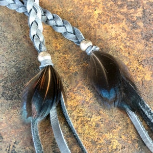May include: A braided leather cord with two black feather tassels and silver metal beads. The cord is gray and the feathers are dark brown.