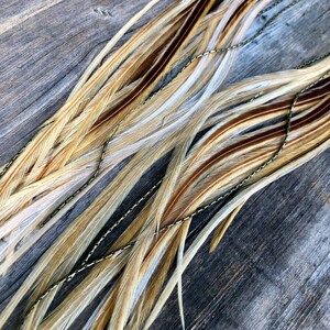 May include: A bundle of long, natural feathers in shades of brown, beige, and white, with a gold chain running through them. The feathers are arranged in a fan shape, with the tips of the feathers pointing to the right.