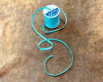 May include: A spool of turquoise-colored flat cord with a matching length of cord unraveled. The cord is on a brown, textured surface. The spool has a white label with blue text.
