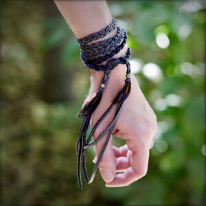 May include: A dark gray leather bracelet with feather accents and metal beads. The bracelet is wrapped around a wrist.