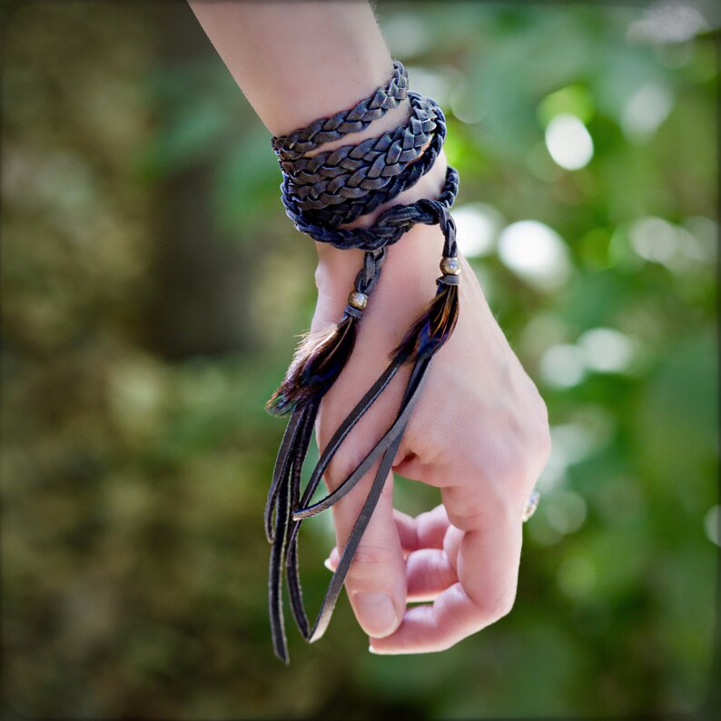 May include: A dark gray leather bracelet with feather accents and metal beads. The bracelet is wrapped around a wrist.