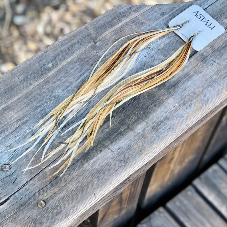 May include: A pair of long, feather earrings with a gold chain detail. The feathers are a mix of white, brown, and beige. The earrings are labeled "ASTALI" on a white card.