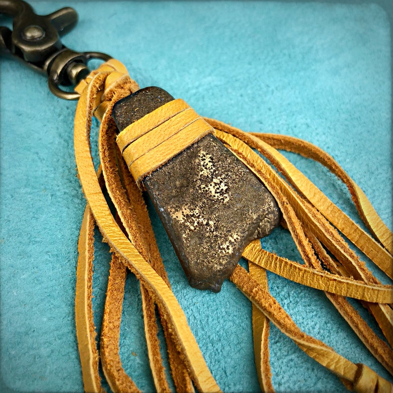 May include: A brown leather tassel keychain with a gold-toned metal clasp and a brown stone pendant. The tassel is made of multiple strands of leather and has a frayed edge.