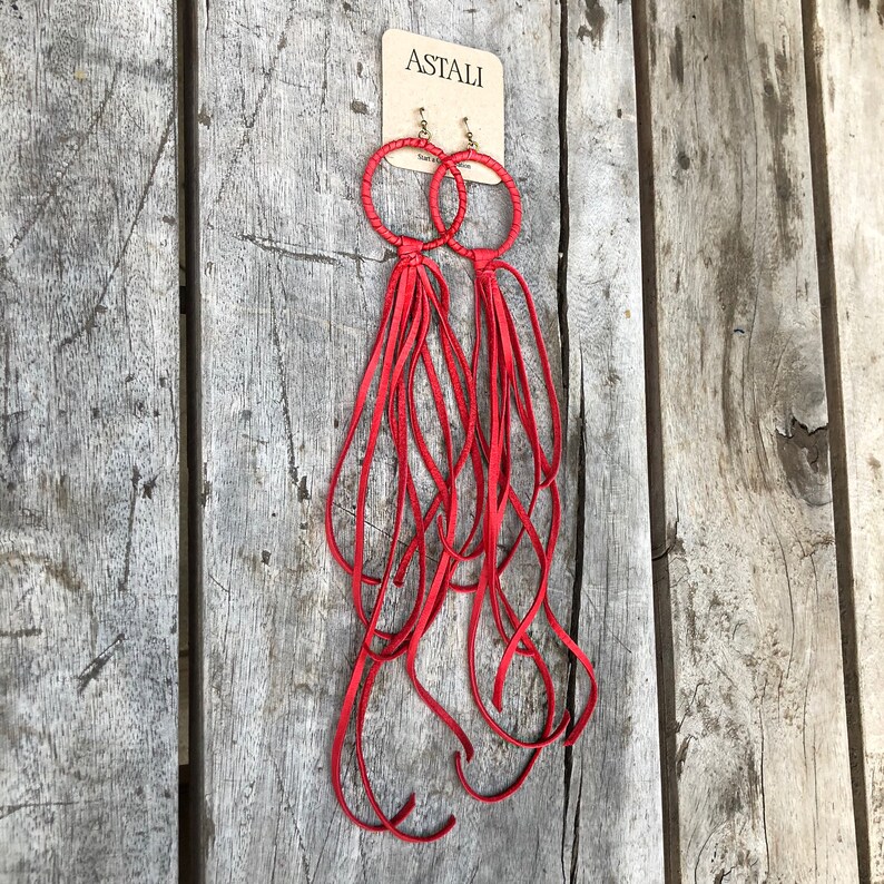 May include: Red leather earrings with a hoop design and long fringe tassels. The earrings are labelled "ASTALI" on a white tag.