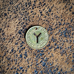May include: A vintage brass token with the text "GOOD FOR ONE FARE" and "NYC" in the center. The token has a hole at the top and is set against a textured brown background with dark blue speckles.