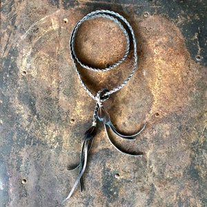 May include: A black and white braided leather necklace with a feather tassel. The necklace is on a rustic metal background.