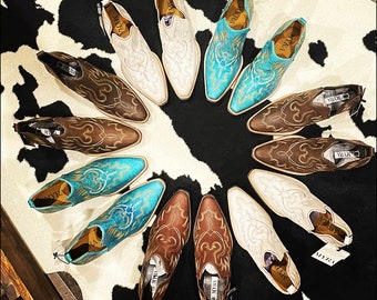 May include: A collection of 12 brown and turquoise ankle boots with intricate embroidery and pointed toes. The boots are arranged in a circle on a white and black cowhide rug.
