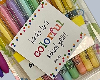 May include: A set of colorful pens with a gift tag that reads "Here's to a colorful school year!"  The pens are packaged in a clear plastic container with a yellow and orange background. The gift tag is white with black text and colorful polka dots.