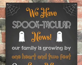 Op de afbeelding: Een zwart schoolbordbord met witte tekst die aankondigt "We Have Spook-tacular News! Our family is growing by one heart and two feet. Our Little Monster is due July 2021." Het bord is versierd met twee witte geesten en een rij oranje pompoenen.