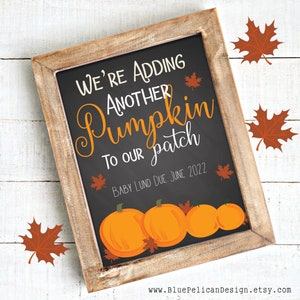 May include: A chalkboard sign with a wooden frame announcing a pregnancy with the text "We're adding another pumpkin to our patch. Baby [Last Name] due June 2022". The sign is decorated with orange pumpkins and fall leaves.