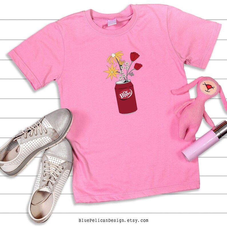 May include: Pink t-shirt with a graphic of a Dr Pepper can with flowers coming out of the top. The flowers are yellow, pink, and white.