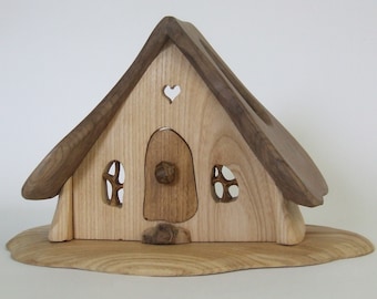 May include: Wooden fairy house with a light brown body and a darker brown roof. The house features a heart-shaped cutout, two small windows, and a rounded door. The base is a light brown, organic shape.