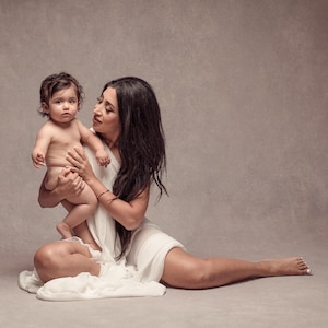 以下が含まれることがあります： 白いドレスを着た女性が赤ちゃんを抱っこしている。女性は足を伸ばして床に座っている。赤ちゃんは女性を見ている。