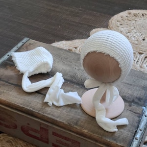 May include: White crocheted baby hat with a wooden doll head and stand. The hat is made of a soft, stretchy fabric and has a simple, elegant design.