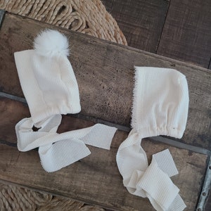 May include: Two white waffle knit baby bonnets with white pom poms and ties. The bonnets are on a wooden surface.