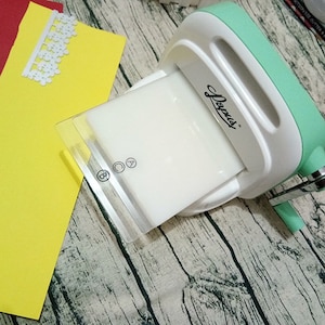 Puede incluir: Una m&aacute;quina de estampar de pl&aacute;stico blanco y verde con una placa de pl&aacute;stico transparente. La m&aacute;quina tiene un mango y una palanca. La m&aacute;quina est&aacute; sobre una superficie de madera.