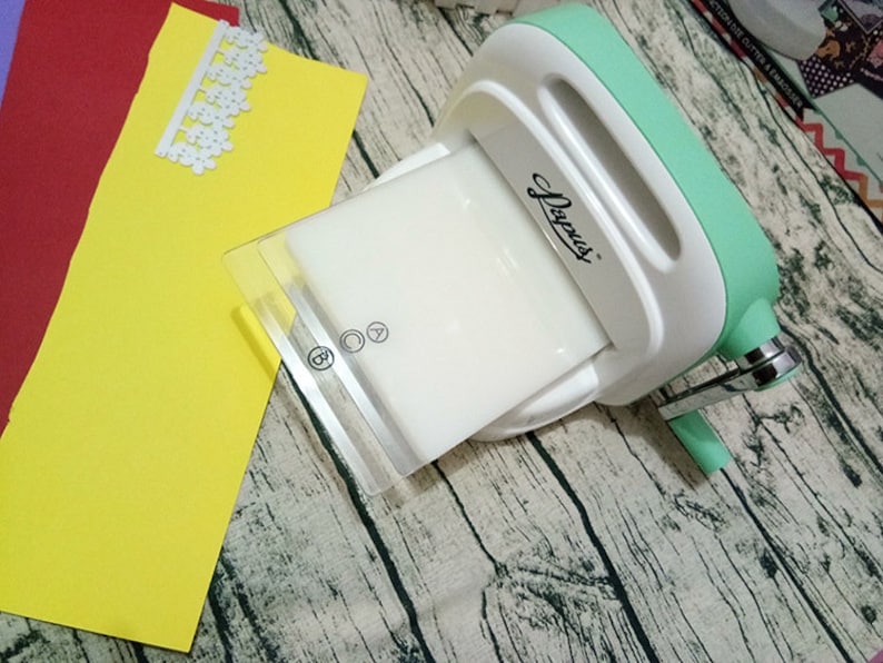 Puede incluir: Una m&aacute;quina de estampar de pl&aacute;stico blanco y verde con una placa de pl&aacute;stico transparente. La m&aacute;quina tiene un mango y una palanca. La m&aacute;quina est&aacute; sobre una superficie de madera.