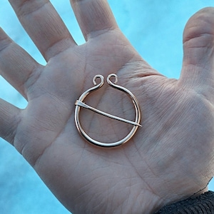 May include: A gold-toned metal ring brooch with a decorative pin. The brooch is circular with a simple design and a smooth finish.