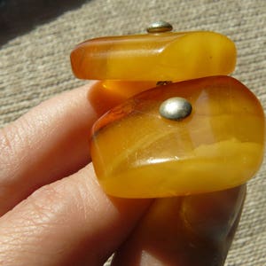 May include: Two pieces of amber jewelry with silver accents. The amber is a warm, honey color and is shaped like a rectangle. The jewelry is on a person's hand.