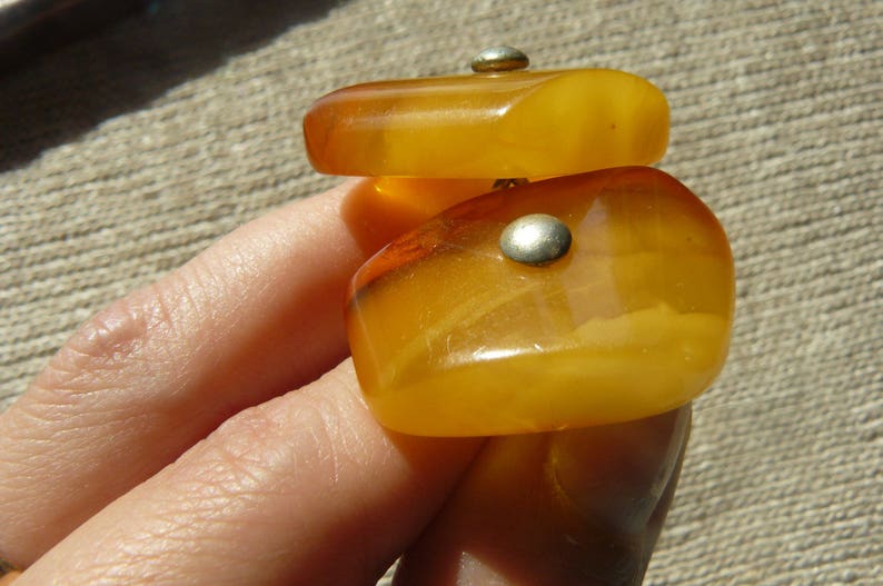 May include: Two pieces of amber jewelry with silver accents. The amber is a warm, honey color and is shaped like a rectangle. The jewelry is on a person's hand.