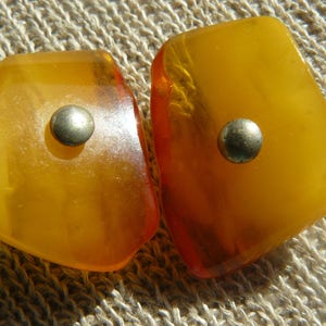 May include: Two amber-colored, irregularly shaped, polished stones with silver metal accents. The stones are set on a textured, beige fabric background.