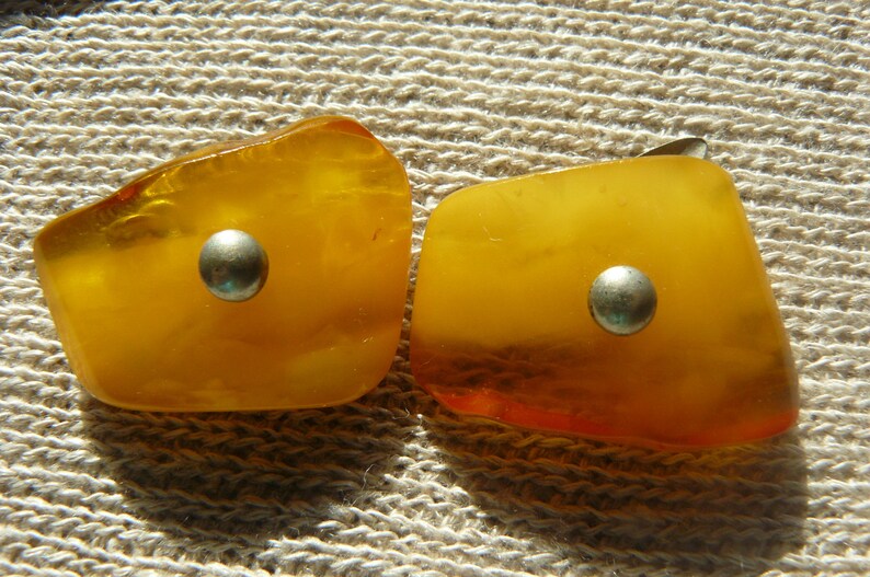 May include: Two amber-colored earrings with silver accents. Each earring has a round, silver-colored metal piece in the center.