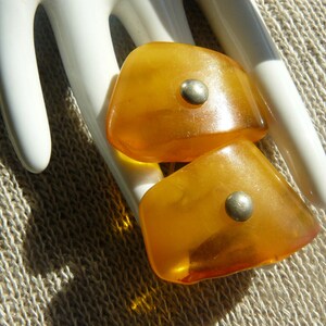 May include: Two amber-colored glass buttons with silver accents. The buttons are shaped like a teardrop and have a smooth, polished finish.