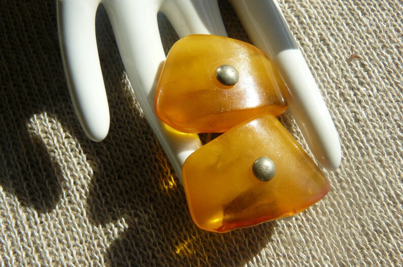 May include: Two amber-colored glass buttons with silver accents. The buttons are shaped like a teardrop and have a smooth, polished finish.